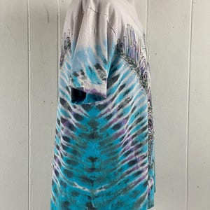 Puede incluir: Una camiseta tie-dye con un esquema de color azul, morado y blanco. La camiseta presenta un gr&aacute;fico de una ciudad de cristal con una monta&ntilde;a en el fondo.