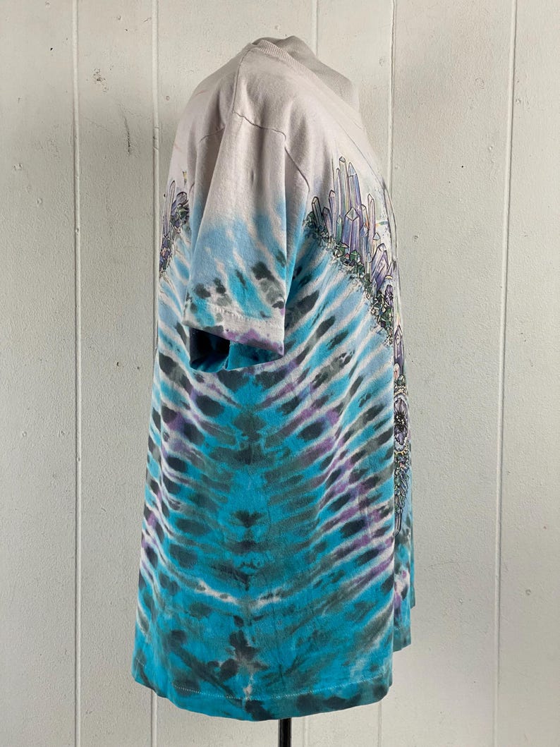 Puede incluir: Una camiseta tie-dye con un esquema de color azul, morado y blanco. La camiseta presenta un gr&aacute;fico de una ciudad de cristal con una monta&ntilde;a en el fondo.