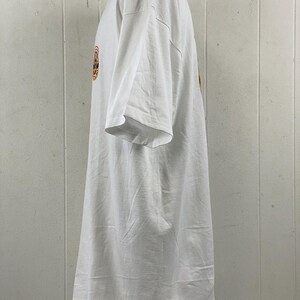 May include: White t-shirt with a short sleeve design. The shirt has a round graphic on the front with orange, yellow, and blue colors. The shirt is made of a soft, comfortable material.