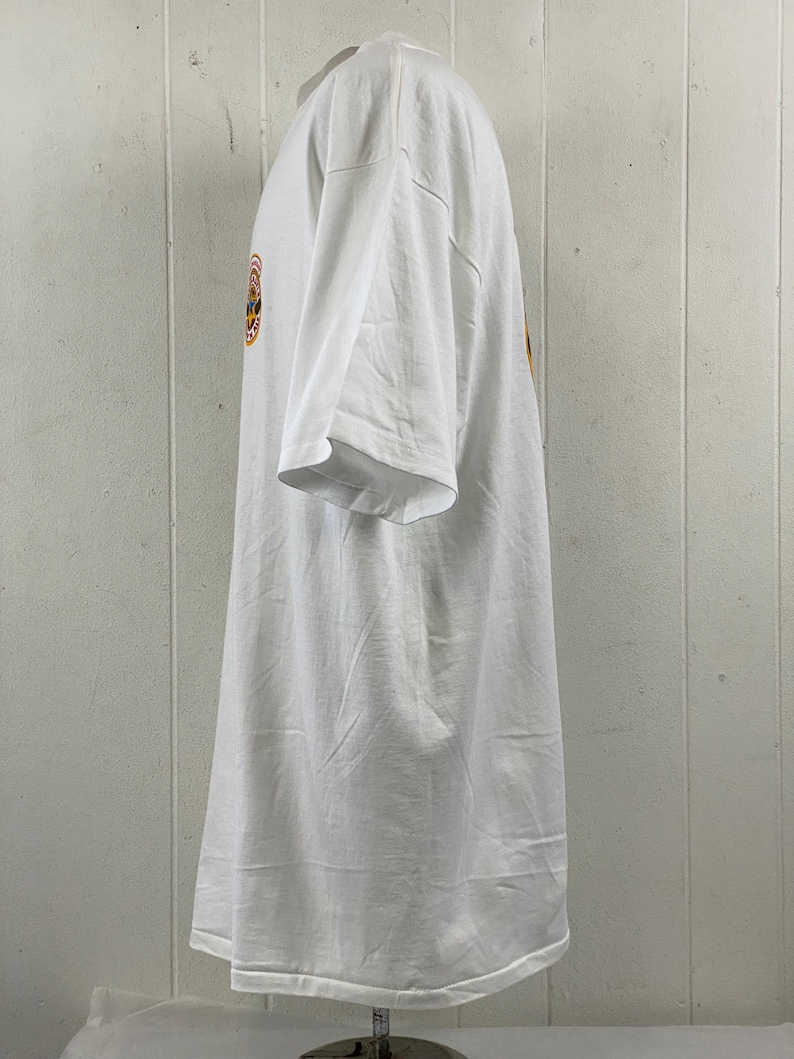 May include: White t-shirt with a short sleeve design. The shirt has a round graphic on the front with orange, yellow, and blue colors. The shirt is made of a soft, comfortable material.