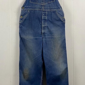K&ouml;nnte beinhalten: Vintage blaue Jeans-Latzhose mit verstellbaren Tr&auml;gern und Knopfverschl&uuml;ssen. Die Latzhose weist Gebrauchsspuren auf, mit ausgeblichenen Stellen und Verf&auml;rbungen. Die Latzhose hat Taschen und das Design ist klassische Arbeitskleidung.