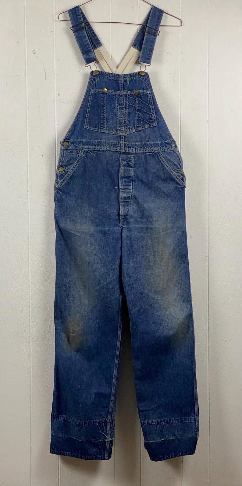 K&ouml;nnte beinhalten: Vintage blaue Jeans-Latzhose mit verstellbaren Tr&auml;gern und Knopfverschl&uuml;ssen. Die Latzhose weist Gebrauchsspuren auf, mit ausgeblichenen Stellen und Verf&auml;rbungen. Die Latzhose hat Taschen und das Design ist klassische Arbeitskleidung.