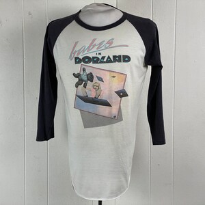 Puede incluir: Una camiseta de estilo béisbol blanca y negra con un gráfico de una mujer y un osito de peluche volando en el aire. El texto "Babies in Borland" está impreso en la camiseta.