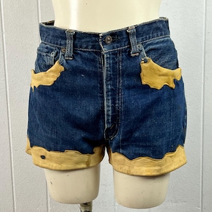 May include: Dark blue denim shorts featuring tan leather trim along the front pockets and the bottom hem. The shorts have a button fly closure, belt loops, and a classic five-pocket design with a visible metal button at the waist.