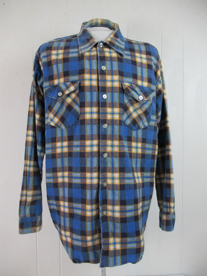 以下が含まれることがあります： 青、黄、茶色の格子柄のフランネルシャツ。長袖でボタン留めです。