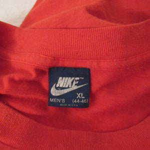 Puede incluir: Una camiseta roja con una etiqueta azul de Nike que dice "Nike" y "Men's XL (44-46)" y "Made in U.S.A."