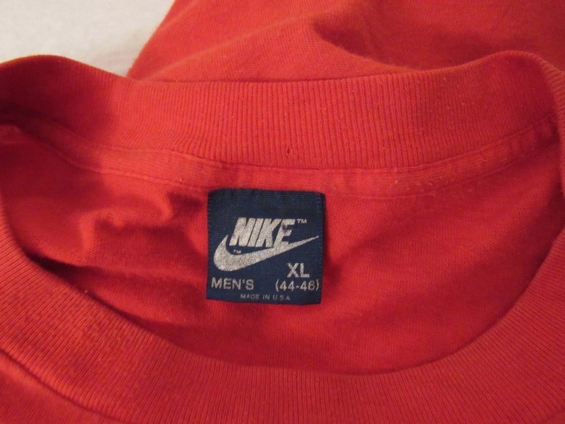 Puede incluir: Una camiseta roja con una etiqueta azul de Nike que dice "Nike" y "Men's XL (44-46)" y "Made in U.S.A."