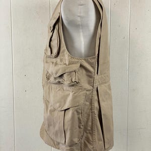 May include: A tan-colored vest with multiple pockets of varying sizes. The vest is displayed on a mannequin and appears to be made of a durable fabric. The vest has a classic design, suitable for outdoor activities or work.