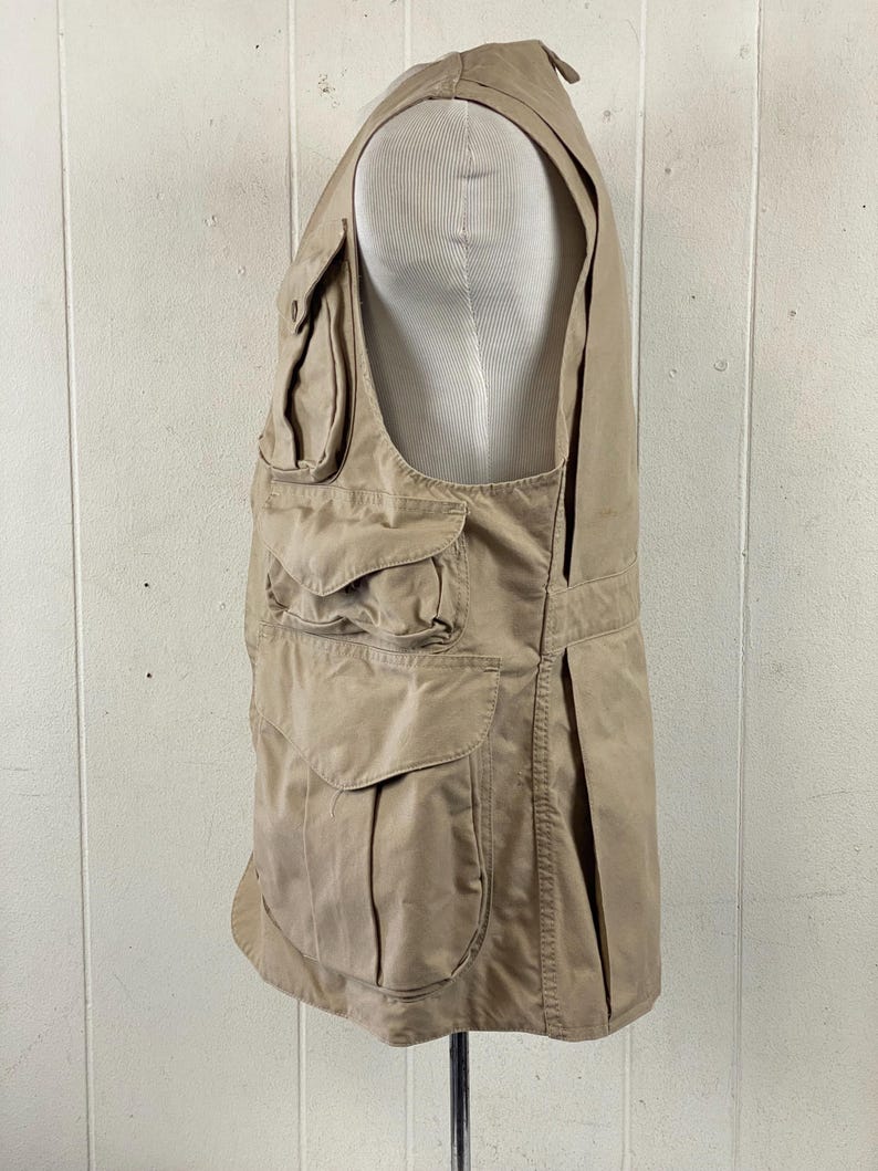 May include: A tan-colored vest with multiple pockets of varying sizes. The vest is displayed on a mannequin and appears to be made of a durable fabric. The vest has a classic design, suitable for outdoor activities or work.
