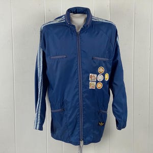 May include: A vintage navy blue Adidas track jacket with a full zipper, light blue stripes on the sleeves, and several patches. The jacket has a stand-up collar and two front pockets. The Adidas logo is on the lower right.