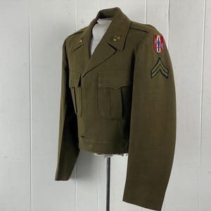 May include: A green military jacket with a patch on the sleeve and a rank insignia. The jacket has a button-up front and two pockets.
