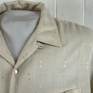 May include: A beige short-sleeved shirt featuring a subtle white diamond-shaped embroidery pattern throughout. The shirt has a classic pointed collar, a button-down front with a single visible white button, and a structured fit on a mannequin.
