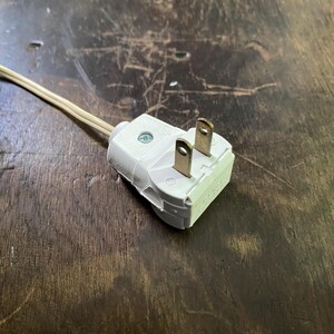 May include: A white electrical plug with two prongs and a beige cable. The plug is made of plastic and has a screw. The cable is thin and flexible. The plug is on a dark wooden surface.