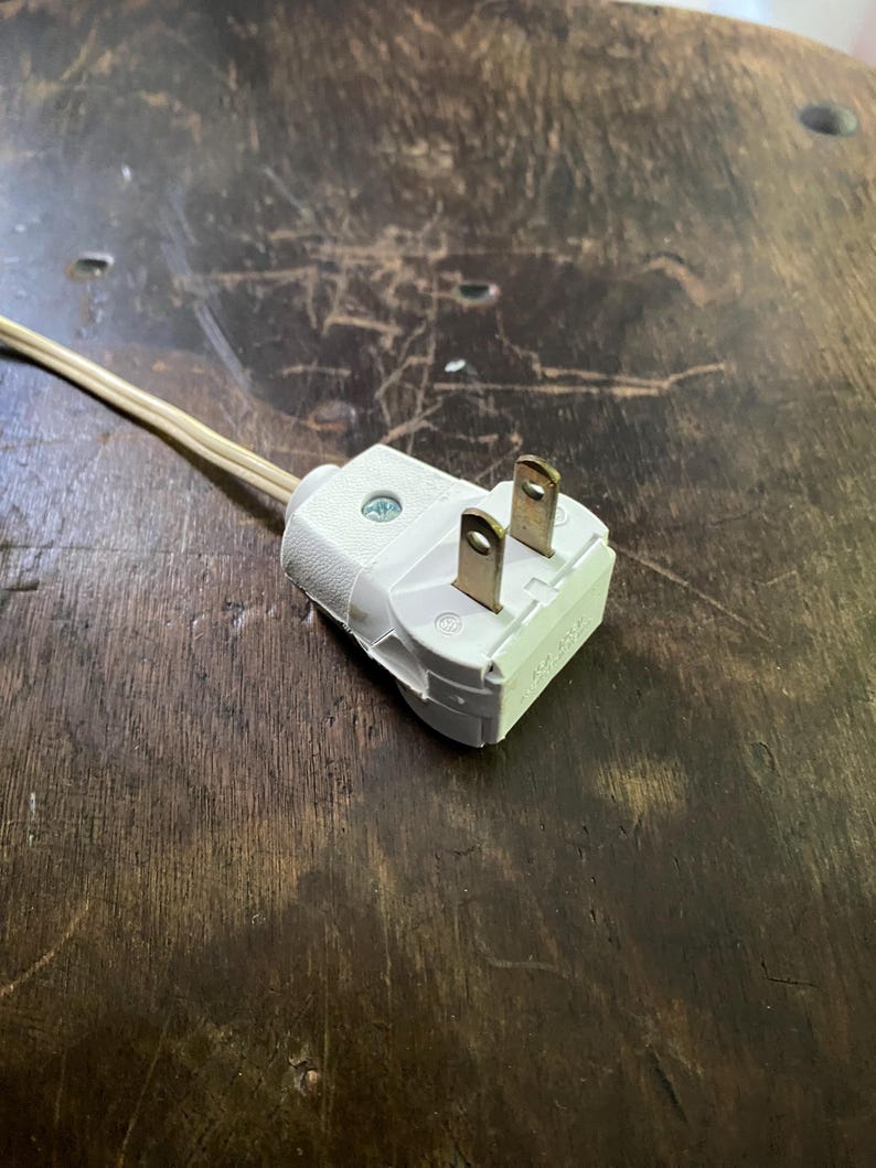 May include: A white electrical plug with two prongs and a beige cable. The plug is made of plastic and has a screw. The cable is thin and flexible. The plug is on a dark wooden surface.