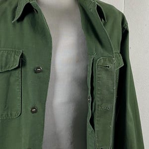 May include: A dark green jacket with a collared neckline and button-down closure. The jacket has two front pockets and a chest pocket. The jacket is displayed on a mannequin. The jacket is made of a durable fabric.