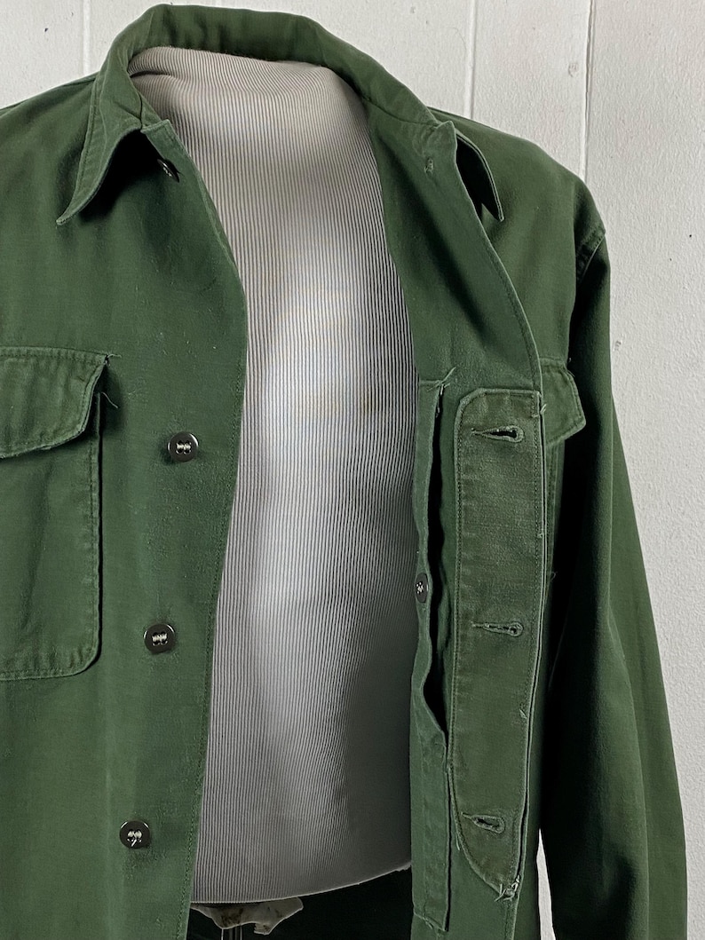 May include: A dark green jacket with a collared neckline and button-down closure. The jacket has two front pockets and a chest pocket. The jacket is displayed on a mannequin. The jacket is made of a durable fabric.