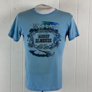 K&ouml;nnte beinhalten: Hellblaues T-Shirt mit einem grafischen Aufdruck einer Bergszene und dem Text "Banff Alberta".