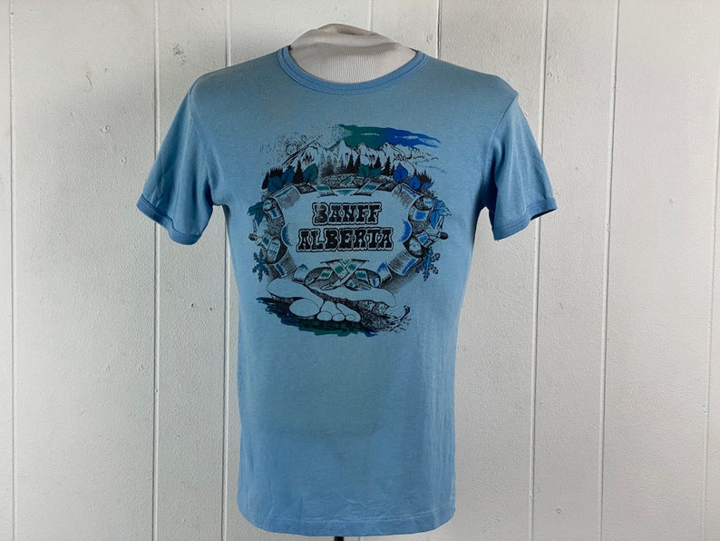K&ouml;nnte beinhalten: Hellblaues T-Shirt mit einem grafischen Aufdruck einer Bergszene und dem Text "Banff Alberta".