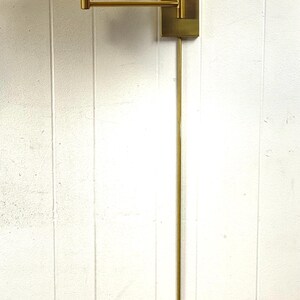 May include: A wall-mounted lamp with a brass finish. The lamp has a rectangular base, an adjustable arm, and a cream-colored lampshade. The lamp's overall height is approximately 122 cm.