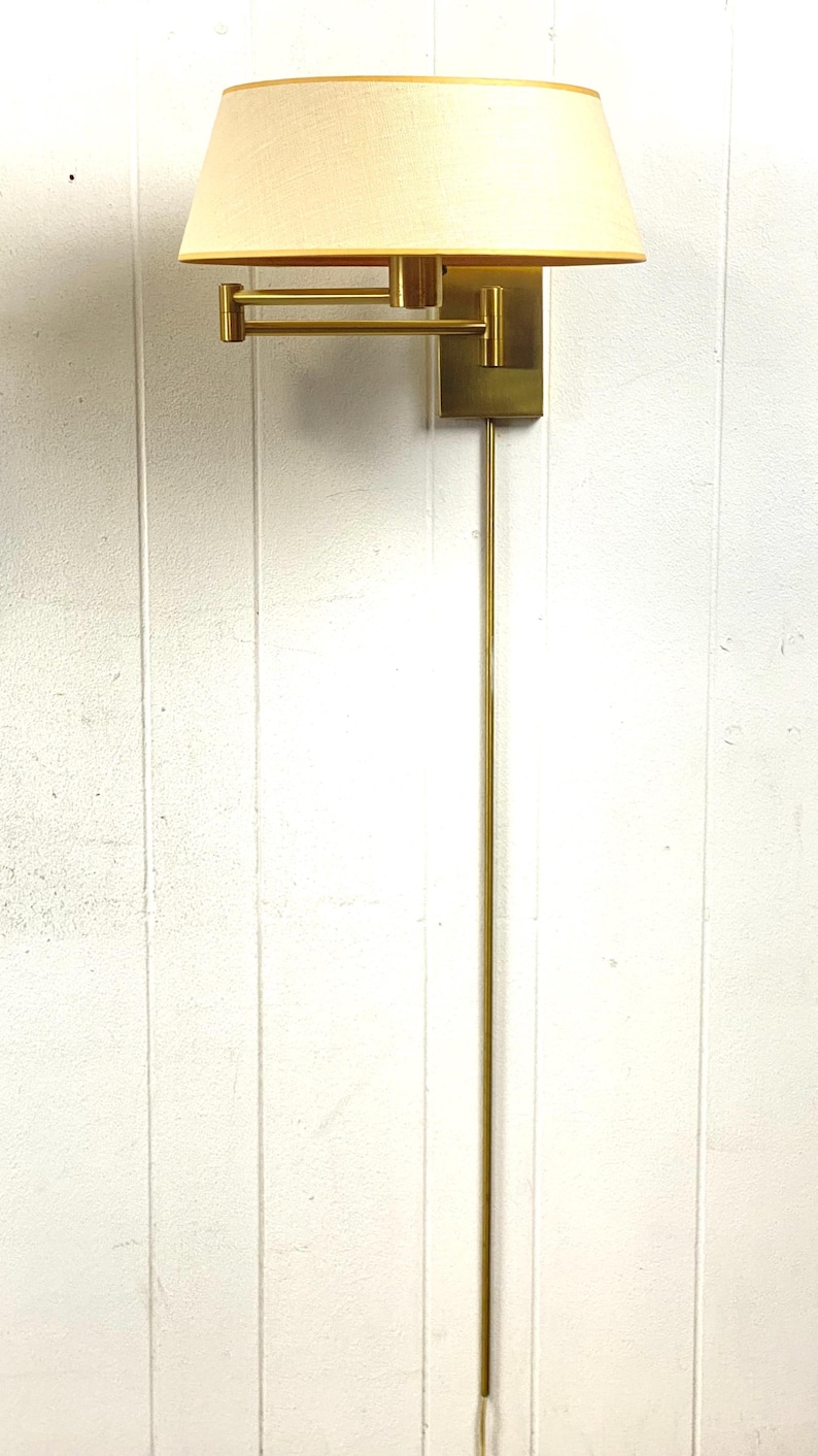 May include: A wall-mounted lamp with a brass finish. The lamp has a rectangular base, an adjustable arm, and a cream-colored lampshade. The lamp's overall height is approximately 122 cm.