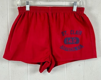 May include: Bright red athletic shorts featuring an elastic waistband and a dark blue graphic print on the right leg. The print displays the text ST. CLAIR above the number 149 inside an oval shape, with the words UNIVERSAL E.D. below the oval.