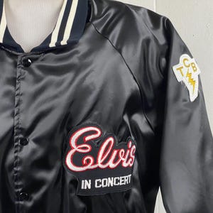 Puede incluir: Chaqueta de satén negro con cuello a rayas blancas y azul marino. Cuenta con un parche "Elvis IN CONCERT" en rojo y blanco, y un parche de rayo "TCB". La chaqueta tiene botones negros.