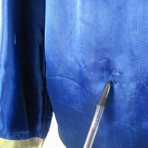 May include: A close-up of a blue satin fabric with a small hole in the center. A black pen is pointing at the hole.