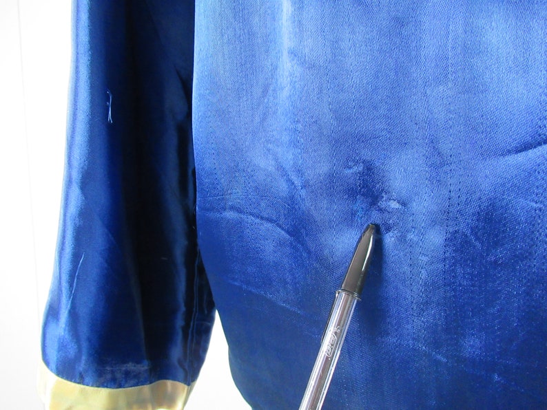 May include: A close-up of a blue satin fabric with a small hole in the center. A black pen is pointing at the hole.