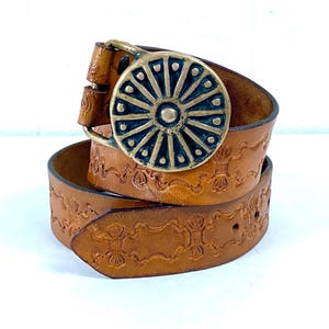May include: A brown leather belt with a decorative brass buckle. The buckle is circular with a sunburst design. The belt has embossed floral patterns and is rolled up. The belt is approximately 1.5 inches wide.