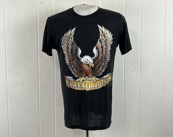 May include: A black short-sleeve t-shirt featuring a graphic of a bald eagle with spread wings perched on a stone banner. The text Harley-Davidson is printed in yellow and white lettering across the stone banner below the eagle illustration.