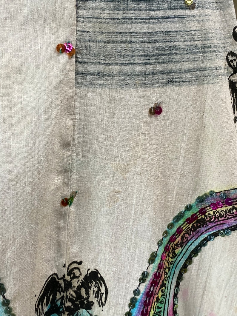 May include: A white linen fabric with a colorful rainbow design and sequins. The fabric has a black and white striped pattern in the upper left corner. There are several sequins in various colors, including pink, green, and gold.