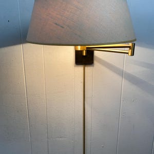 May include: A wall-mounted lamp with a beige lampshade and a gold-coloured frame. The lamp has a rectangular black base and a long, slender gold-coloured stem. The lampshade is oval-shaped and has a gold trim.