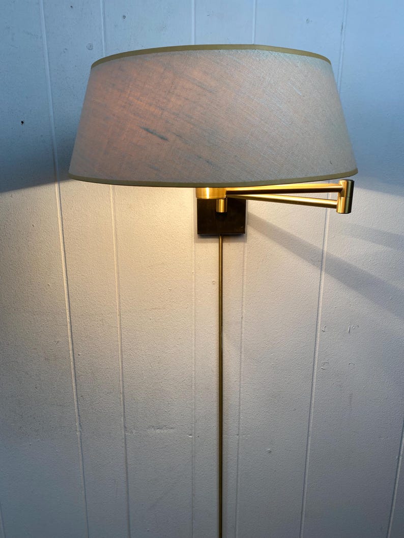 May include: A wall-mounted lamp with a beige lampshade and a gold-coloured frame. The lamp has a rectangular black base and a long, slender gold-coloured stem. The lampshade is oval-shaped and has a gold trim.