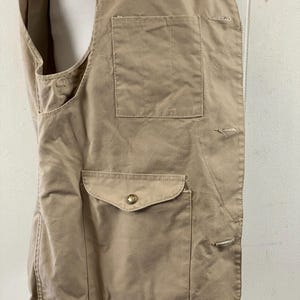 May include: Tan-colored vest with two front pockets and a button closure. The vest has a tailored fit and is made of a durable fabric. The pockets are rectangular and have a flap closure. The vest is designed for outdoor activities.