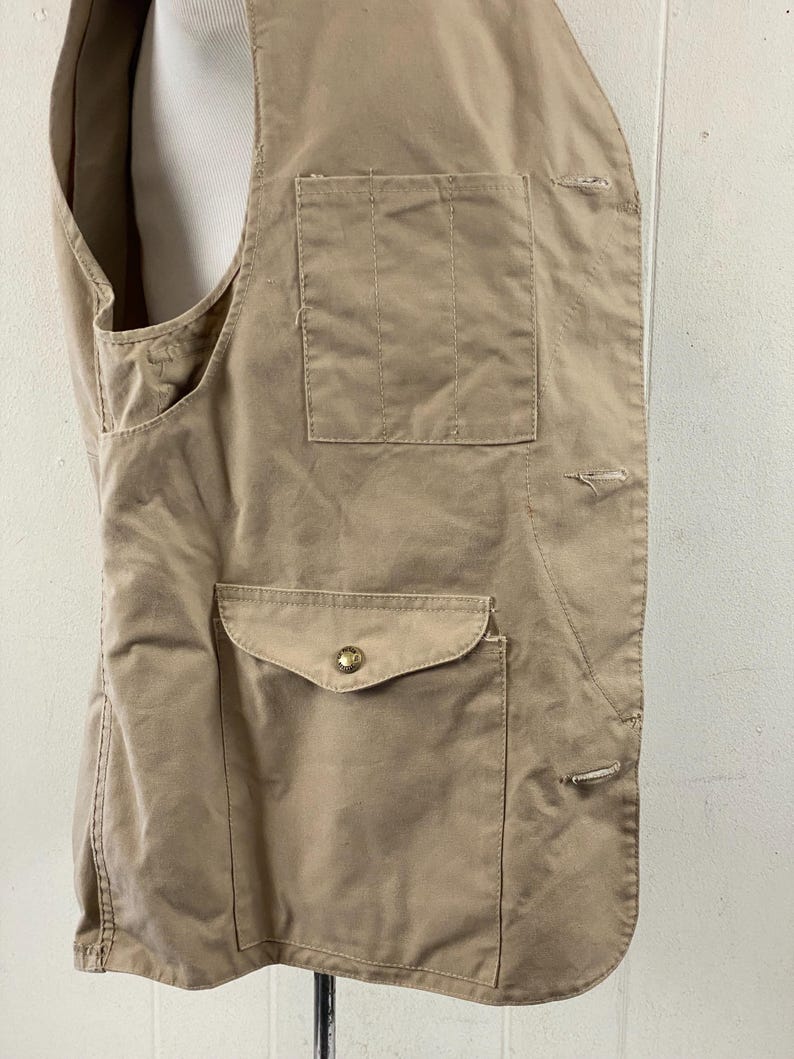 May include: Tan-colored vest with two front pockets and a button closure. The vest has a tailored fit and is made of a durable fabric. The pockets are rectangular and have a flap closure. The vest is designed for outdoor activities.