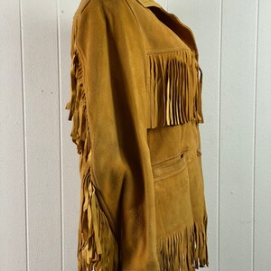 Puede incluir: Una chaqueta de gamuza marr&oacute;n con detalles de flecos en las mangas, el dobladillo y el cuello. La chaqueta tiene cierre de botones y un bolsillo en la parte delantera.