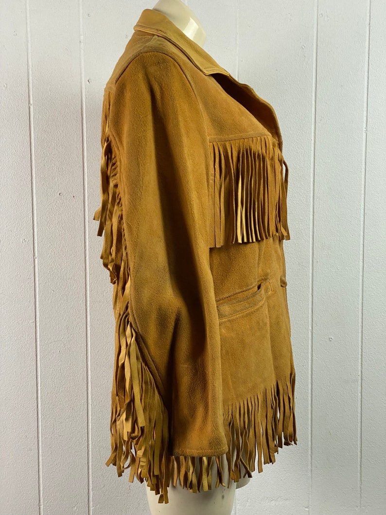 Puede incluir: Una chaqueta de gamuza marr&oacute;n con detalles de flecos en las mangas, el dobladillo y el cuello. La chaqueta tiene cierre de botones y un bolsillo en la parte delantera.