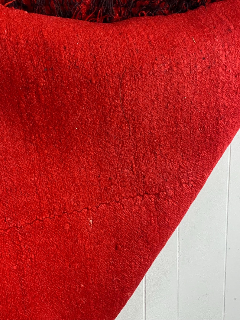 May include: A close-up of a red woven rug with a fringe border. The rug is folded in half and the texture of the fabric is visible.