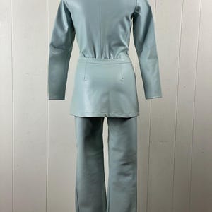 K&ouml;nnte beinhalten: Ein hellblauer Kunstleder-Jumpsuit mit Stehkragen und langen &Auml;rmeln. Der Jumpsuit hat einen Rock und eine Hose.