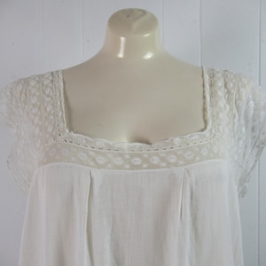 Puede incluir: Camis&oacute;n blanco de algod&oacute;n con ribete de encaje alrededor del escote y las mangas. El camis&oacute;n tiene un escote cuadrado y un corpi&ntilde;o fruncido.