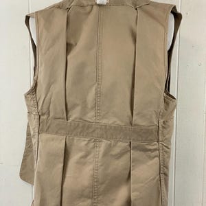 May include: A tan-colored vest with multiple pockets and a tailored fit. The back of the vest features vertical pleats and a horizontal band across the lower back. The vest is displayed on a mannequin.