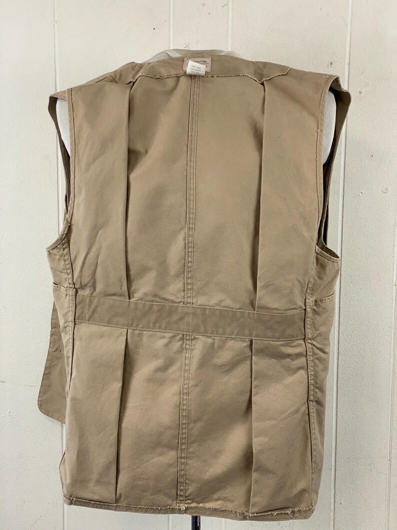 May include: A tan-colored vest with multiple pockets and a tailored fit. The back of the vest features vertical pleats and a horizontal band across the lower back. The vest is displayed on a mannequin.