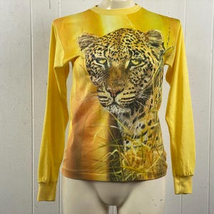 May include: Yellow long-sleeve shirt featuring a detailed leopard face graphic. The shirt has a crew neck and is made of a soft material. The leopard graphic is set against a yellow and brown background, creating a striking visual.