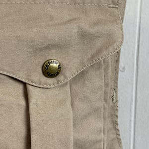 May include: Close-up of a tan-colored garment pocket with a flap and a brass button. The button is embossed with the words "FILSON CO. SEATTLE". The fabric has visible stitching and a slightly worn appearance.