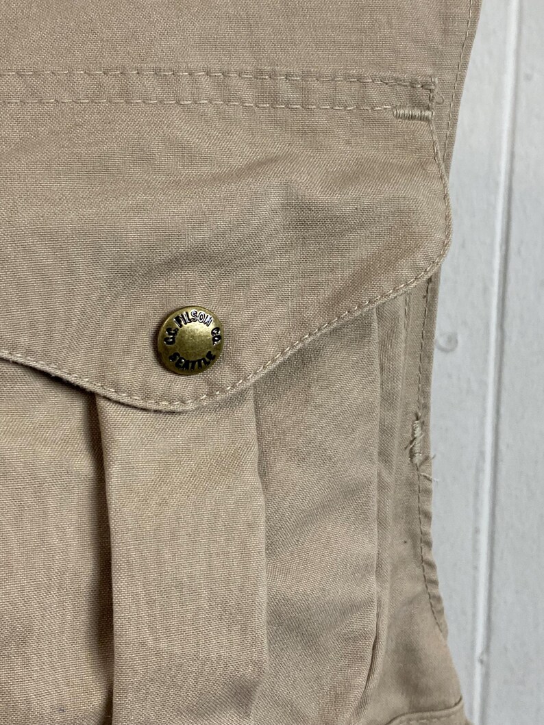May include: Close-up of a tan-colored garment pocket with a flap and a brass button. The button is embossed with the words "FILSON CO. SEATTLE". The fabric has visible stitching and a slightly worn appearance.