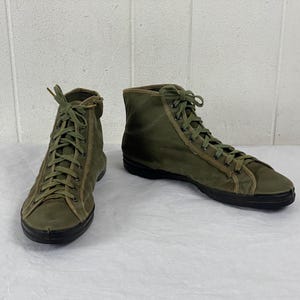 May include: A pair of olive green high-top sneakers with matching laces. The shoes have a canvas-like texture and a black rubber sole. The sneakers are displayed on a white surface.