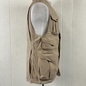 May include: Tan utility vest with multiple pockets of varying sizes. The vest has a button closure and a high collar. The vest is displayed on a mannequin. The vest is made of a durable, lightweight fabric.