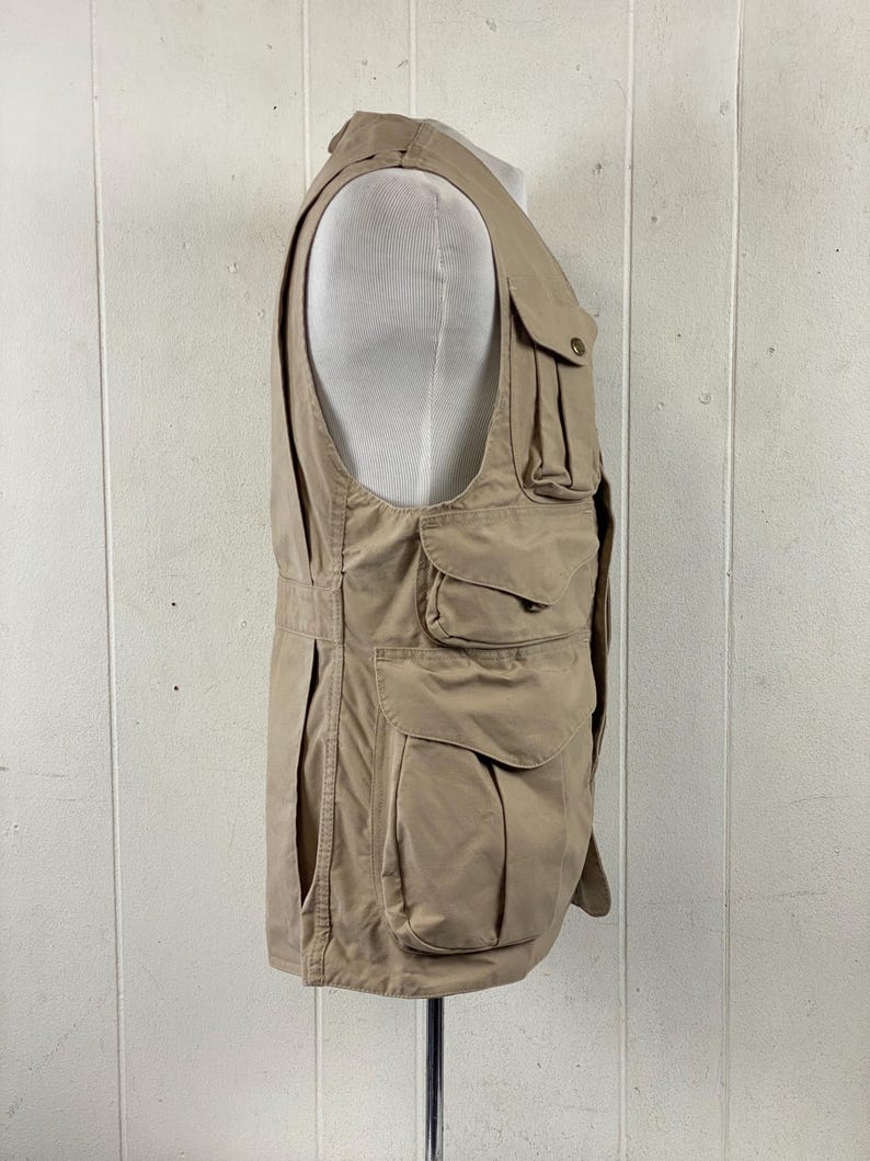 May include: Tan utility vest with multiple pockets of varying sizes. The vest has a button closure and a high collar. The vest is displayed on a mannequin. The vest is made of a durable, lightweight fabric.