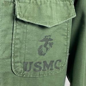 May include: Close-up of a green military shirt with a pocket on the chest. The pocket features a black emblem and the letters "USMC" printed in black. The shirt is made of a durable fabric.