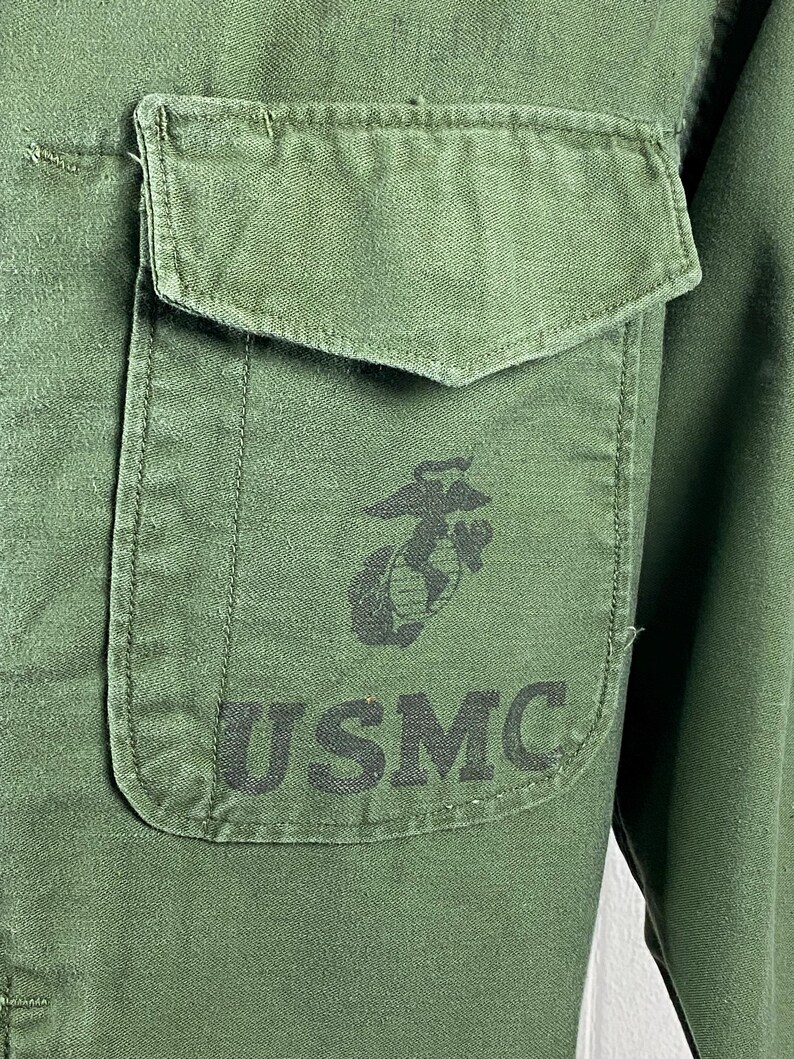 May include: Close-up of a green military shirt with a pocket on the chest. The pocket features a black emblem and the letters "USMC" printed in black. The shirt is made of a durable fabric.